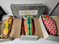 Three boxed silk kites showing yellow, green-orange and red designs in their boxes