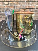 Group view of brass ice bucket with lid, acrylic tray, stainless shaker tin holding strainer, tongs, corkscrew and pourer