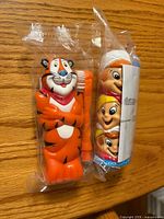 Front view of both sealed toothbrush sets showing Tony the Tiger figure with orange toothbrush and Snap-Crackle-Pop figure with blue toothbrush