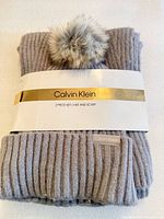 Front view of packaged Calvin Klein hat and scarf set showing light gray rib-knit fabric, faux fur pom, brand band