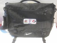 Front view of black Nike messenger bag showing flap, patch, swoosh, strap