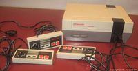 Front and side view of Nintendo Entertainment System console with attached controller cables and three controllers displayed