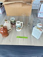 All eight items arranged: two stoneware jugs, Budweiser stein, Munich crest stein, two pilsner glasses, HB glass stein, white eagle decanter