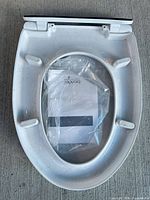 Underside view of Brondell speckled white toilet seat with attached hinge, mounting bracket, and sealed instruction manual