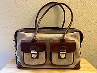 Front view showing wheat canvas body, brown leather trim, double handles, two front flap pockets with nickel clasps