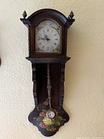 Front view showing entire wooden wall clock with dial, pendulum, floral motif and finials