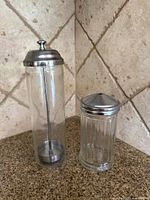 Tall glass straw dispenser and shorter ribbed glass sugar pourer side by side