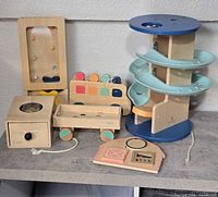 Group shot of all wooden toddler toys including walker cart, pull wagon, spiral ramp tower, ball drop box, activity board and small pieces