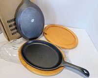 Cast iron skillet resting on wooden trivet, additional skillet and trivet visible, box and packaging included