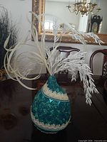 Teal paper mache vase with lace trim holding white faux foliage displayed on table