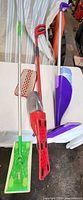 Three long-handled mops (green Swiffer Sweeper, red Vileda spray mop, purple Swiffer WetJet) and small red manual sweeper on table