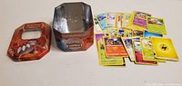 Spread of assorted Pokémon cards beside opened Groudon collector tin showing lid and base