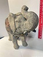 Front three-quarter view of gray resin elephant with raised trunk showing red saddle panel and gold beads