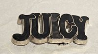 Front view of JUICY belt buckle showing silver frame and black enamel letters