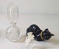 Three perfume bottles: faceted clear crystal with grape stopper, black cat bottle, small ribbed bottle with frosted bird stopper