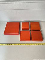 Five closed red-orange wood jewelry boxes beside measuring tape showing relative sizes