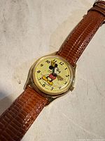 Close view of watch face showing Mickey Mouse graphic, gold-tone case and brown strap texture