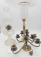 Front view of all three items: glass oil lamp, brass hurricane candlestick, brass multi-arm candelabrum
