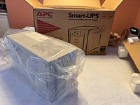 UPS in plastic wrap beside open APC Smart-UPS box with foam inserts