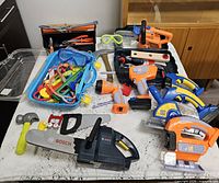 Overall view of assorted plastic toy power tools, hand tools, accessories and Bosch chainsaw on table