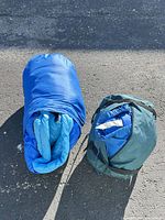 Rolled blue sleeping bag beside dark teal stuff sack on pavement