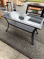 Three-quarter view of coffee table showing glass top, metal frame, woven shelf