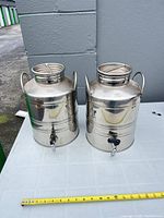 Two stainless 10 L beverage dispensers with spigots and tape measure for scale