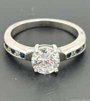 Front angled view of ring showing solitaire clear stone and channel-set blue/clear accents