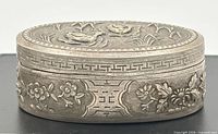 closed silver oval box showing side floral panels and fretwork border