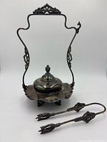 Full view of ornate silver plate pickle castor stand with lid seated in base and tongs in front
