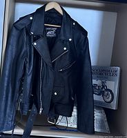 Front view of Event Leather motorcycle jacket on hanger with book and part of metal chest visible