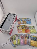 Cardboard storage box filled with large stack of Pokémon reverse holo trading cards plus several cards spread out to show foil surfaces