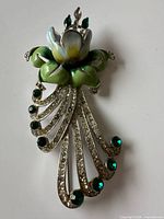 Front view of enamel flower brooch showing painted petals, green leaves, rhinestone scrolls and green stones