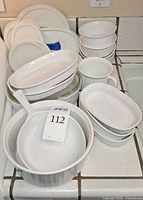 Assortment of white CorningWare French White dishes including oval casseroles, round casseroles, long-handled saucepan, ramekins, mugs and lids stacked on countertop