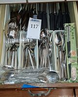 Drawer view showing assorted stainless steel forks, spoons, sealed teaspoon pack, Victorinox steak knives with black handles, serving spoon and packaged chopsticks