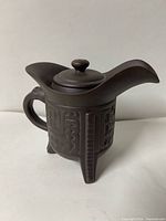 Front view of dark clay teapot on three legs, relief pattern visible