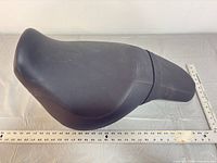 Top view of entire black vinyl motorcycle seat with ruler showing length around 22 in.