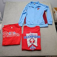 All three Phillies clothing items laid out: blue jacket, two red T-shirts