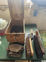 Open wooden shoe shine box with cloths inside, two large brushes, small brush, black polish tin on table