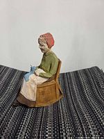 Side view of small hand-carved wooden elderly woman figurine seated on chair showing carving detail and paint colors