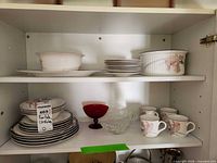Cabinet shelves with stacks of Noritake plates, serving bowls, red glass compote, clear glass bowl