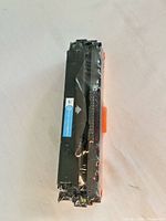 Front view of sealed black toner cartridge with blue compatibility label and orange protective clip