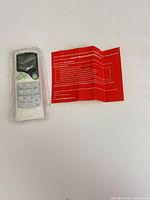 Front view of remote in plastic sleeve beside folded red instruction leaflet