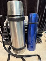 Tall silver stainless steel flask with carry strap beside blue ThermoCafe flask