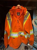 Front view of high-visibility orange safety jacket on hanger showing reflective stripes and hood