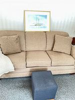 Front view of beige sofa with two pillows, painting resting on back, blue ottoman in foreground