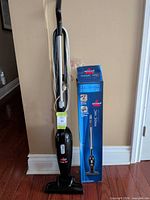 Full view of vacuum standing upright with cord wrapped alongside original box