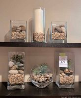 Two shelves holding six glass containers with pebbles, stones, candle, faux plants