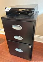 Front view of black metal three-drawer filing cabinet with mesh tray and riser shelf on top
