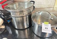 Stock pot with steamer insert and perforated lid beside saucepan with glass lid